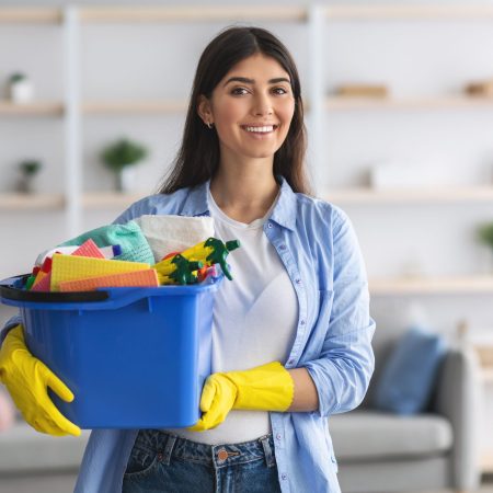Housekeeping Concept. Portrait of smiling young woman holding bucket with cleaning detergents and rags, wearing yellow rubber protective gloves, looking posing at camera standing in modern apartment
