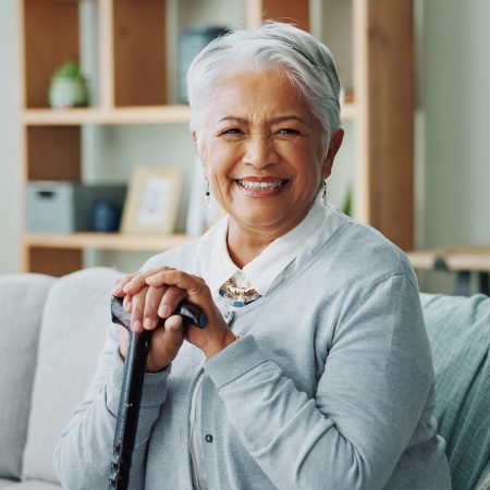 Woman, senior and portrait with cane on sofa, walking support and assistance stick in retirement home on weekend break. Smile, happiness and elderly female person with a disability, wellness and help.
